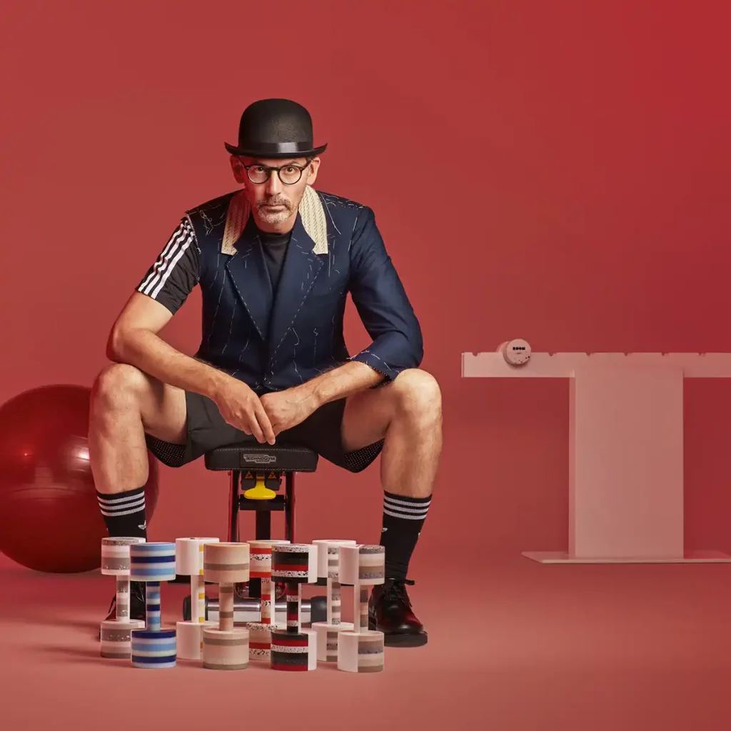 fotografia de jose manuel del estudio de arquitectura Estudi{h}ac posando con los galardones que ha diseñado para room con Krion, en una escena de imitación de haciendo deporte en fondo rojo