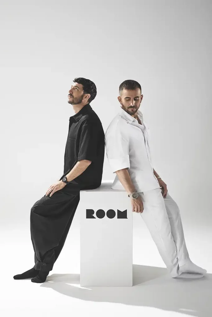 Enfoque de fotógrafo de producto comercial en un estudio minimalista. Dos hombres, uno vestido completamente de blanco y otro de negro, están sentados en cubos blancos apilados. Cada uno sostiene y examina un galardón de diseño cónico minimalista y blanco con líneas de colores. Otros conos de premios similares están dispuestos sobre pedestales en el fondo gris claro.