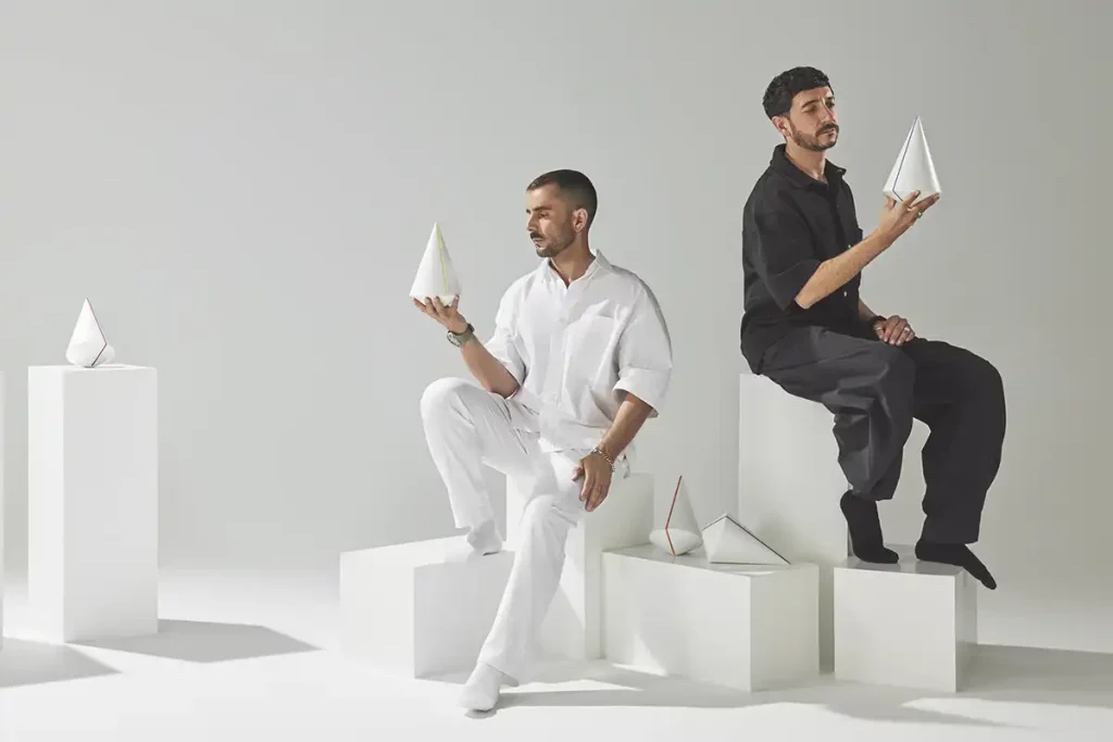 Enfoque de fotógrafo de producto comercial en un estudio minimalista. Dos hombres, uno vestido completamente de blanco y otro de negro, están sentados en cubos blancos apilados. Cada uno sostiene y examina un galardón de diseño cónico minimalista y blanco con líneas de colores. Otros conos de premios similares están dispuestos sobre pedestales en el fondo gris claro.