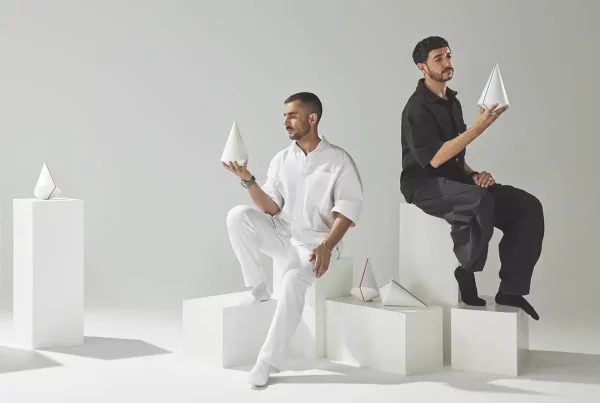 Enfoque de fotógrafo de producto comercial en un estudio minimalista. Dos hombres, uno vestido completamente de blanco y otro de negro, están sentados en cubos blancos apilados. Cada uno sostiene y examina un galardón de diseño cónico minimalista y blanco con líneas de colores. Otros conos de premios similares están dispuestos sobre pedestales en el fondo gris claro.