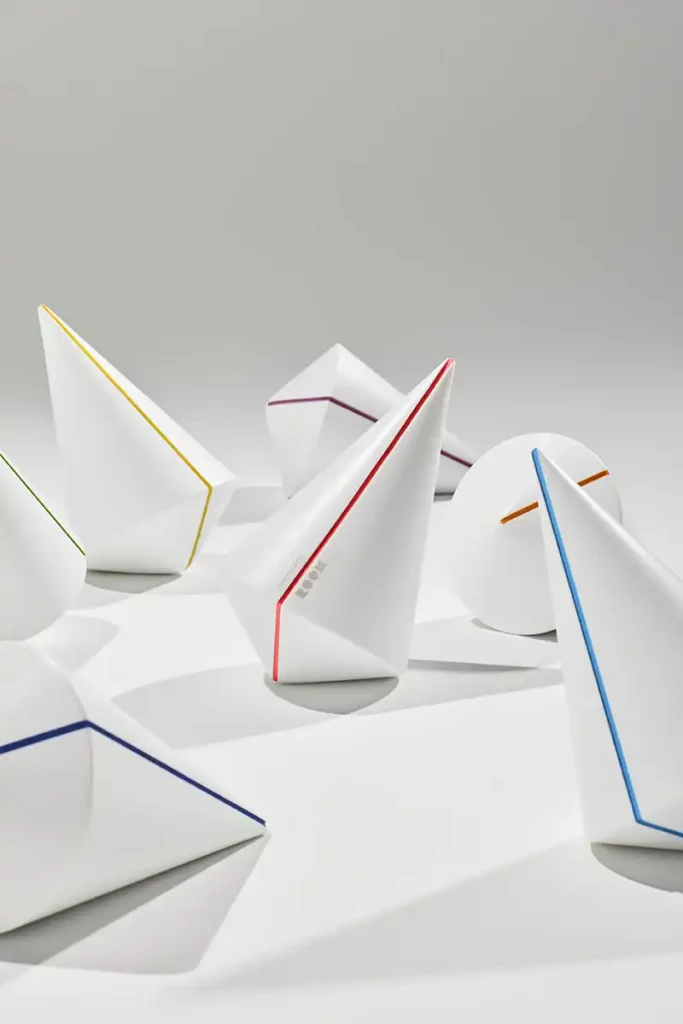 Bodegón de fotografía de producto comercial mostrando varios galardones en forma de peonza para los Premios ROOM. Los trofeos, fabricados en Krion blanco hielo, descansan sobre un fondo neutro y destacan por sus finas líneas grabadas en distintos colores flúor como rojo, azul y amarillo, con el logotipo de la revista sutilmente integrado.