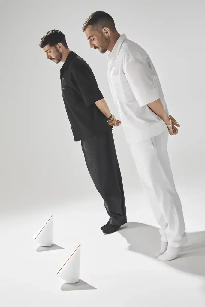 Mut posando uno vestido de blanco y otro de negro, posan en un estudio minimalista rodeados de pedestales blancos. Sobre ellos y en el suelo se exponen los galardones en forma de peonza, fabricados en Krion blanco hielo con líneas en colores flúor. Fotografía de producto comercial de los trofeos para los Premios ROOM. Dos hombres